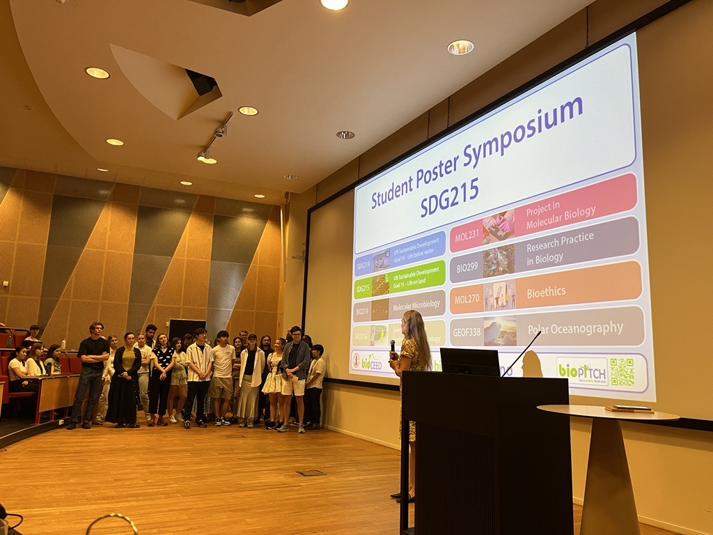 A teacher introduces her course in front of the audience while students are gathered on the stage